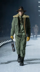 Model walking a snowy runway in an olive parka with shearling collar, brown hat, mustard gloves, and sturdy boots, carrying a water bottle.
