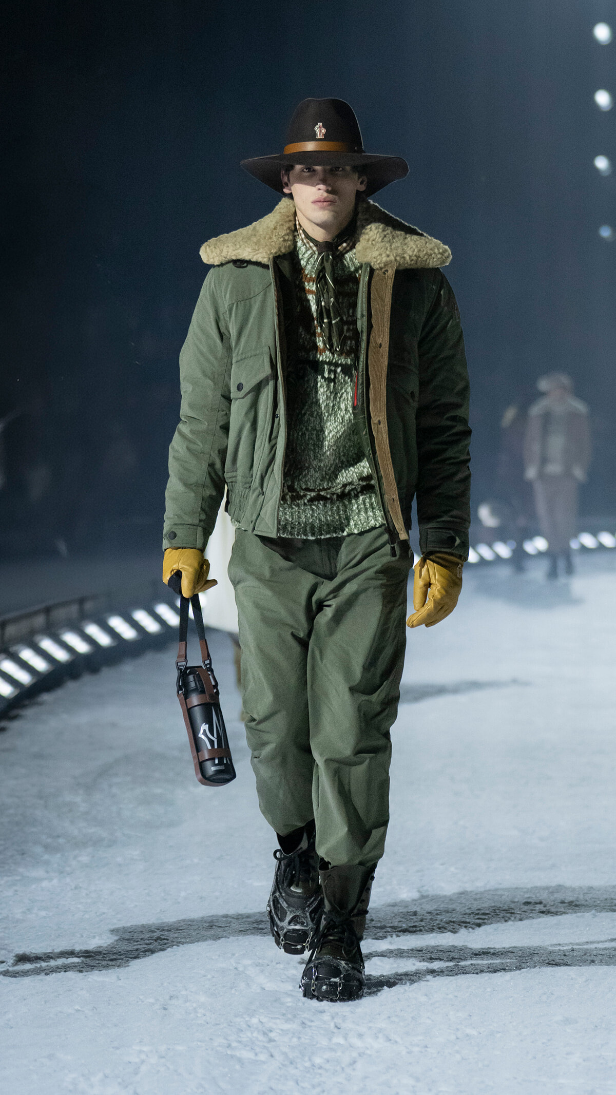 Model walking a snowy runway in an olive parka with shearling collar, brown hat, mustard gloves, and sturdy boots, carrying a water bottle.