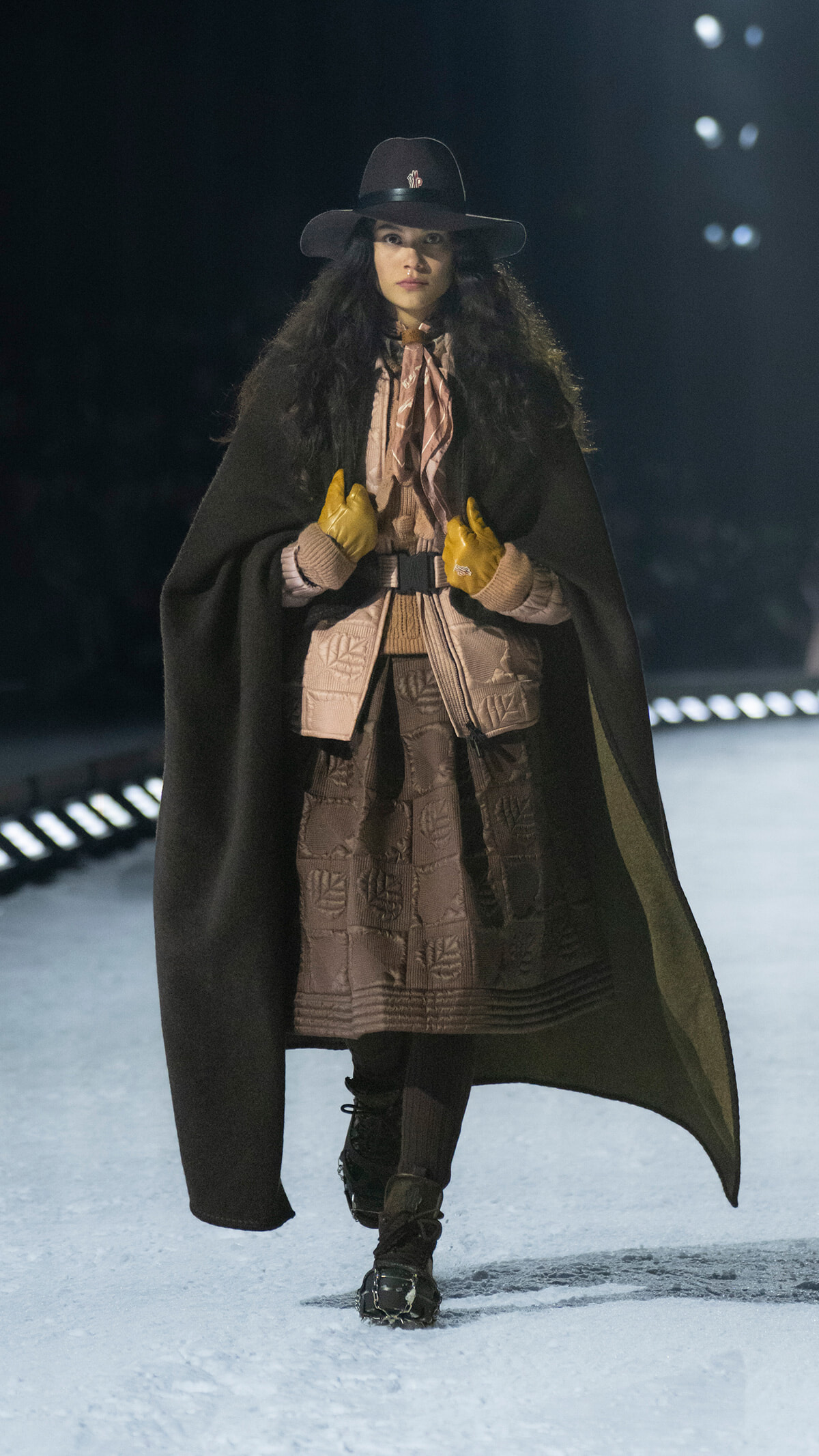 Model on a runway wearing a black cape over a brown quilted dress, wide-brim hat, beige scarf, and yellow gloves.