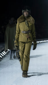 Model walking on a snowy runway in a khaki military-style suit with fur collar and helmet at night fashion show