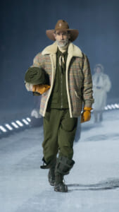 Man walking on an icy runway in a shearling plaid jacket, olive pants, brown hat, holding a rolled blanket and gloves.