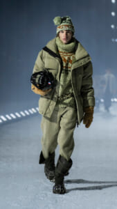 Model in an olive winter coat, chunky knit sweater and hat, walking on an icy runway while holding a helmet.