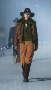 Model walking an icy runway in a brown fur-collared leather jacket, green sweater, tan pants, and a wide-brim black hat.