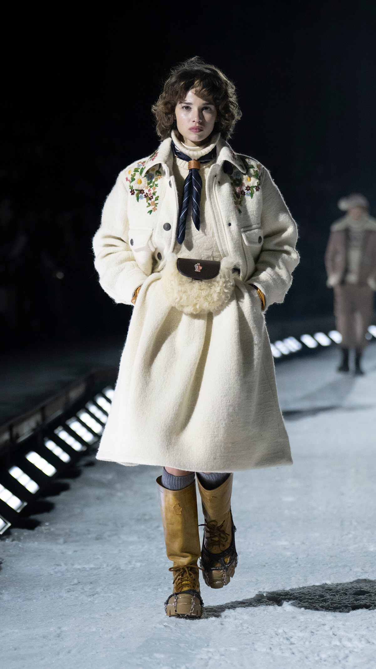 Female model walking a snowy runway in a cream wool coat with floral embroidery, a brown belt bag, and tan boots.