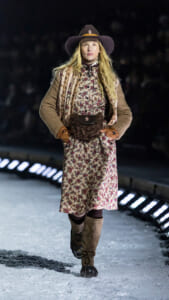 Model walking on a snowy runway at night, wearing a floral dress with a brown scarf, tan cardigan, wide-brim hat, knee-high boots, and a fuzzy belt bag.