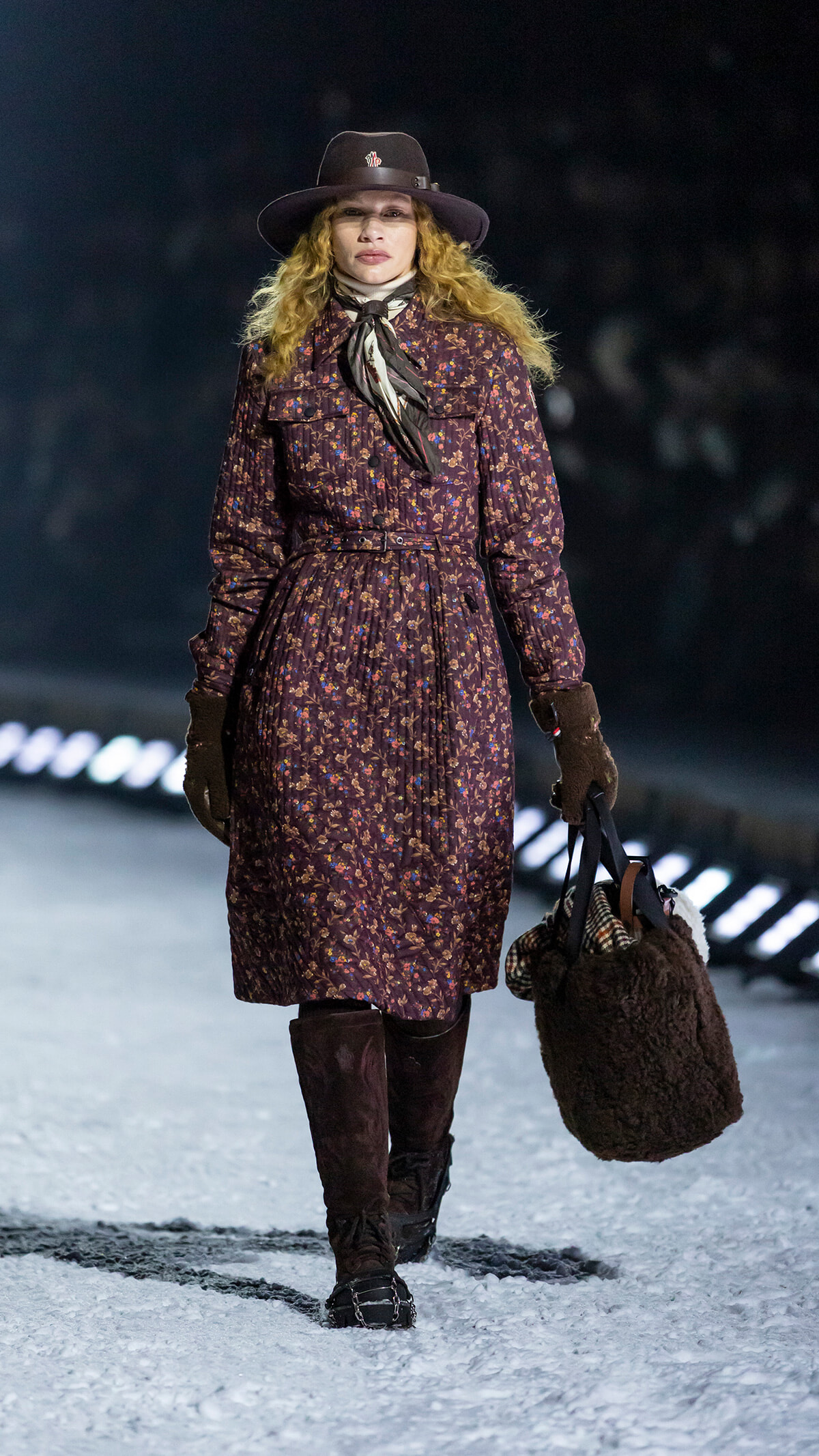 Woman walking in the snow wearing a dark floral coat, brown hat, scarf, gloves, tall boots, and carrying a large brown furry tote bag.