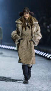 Model walks a snowy runway in a vintage floral dress, oversized fur coat, wide-brim hat, and knee-high boots.