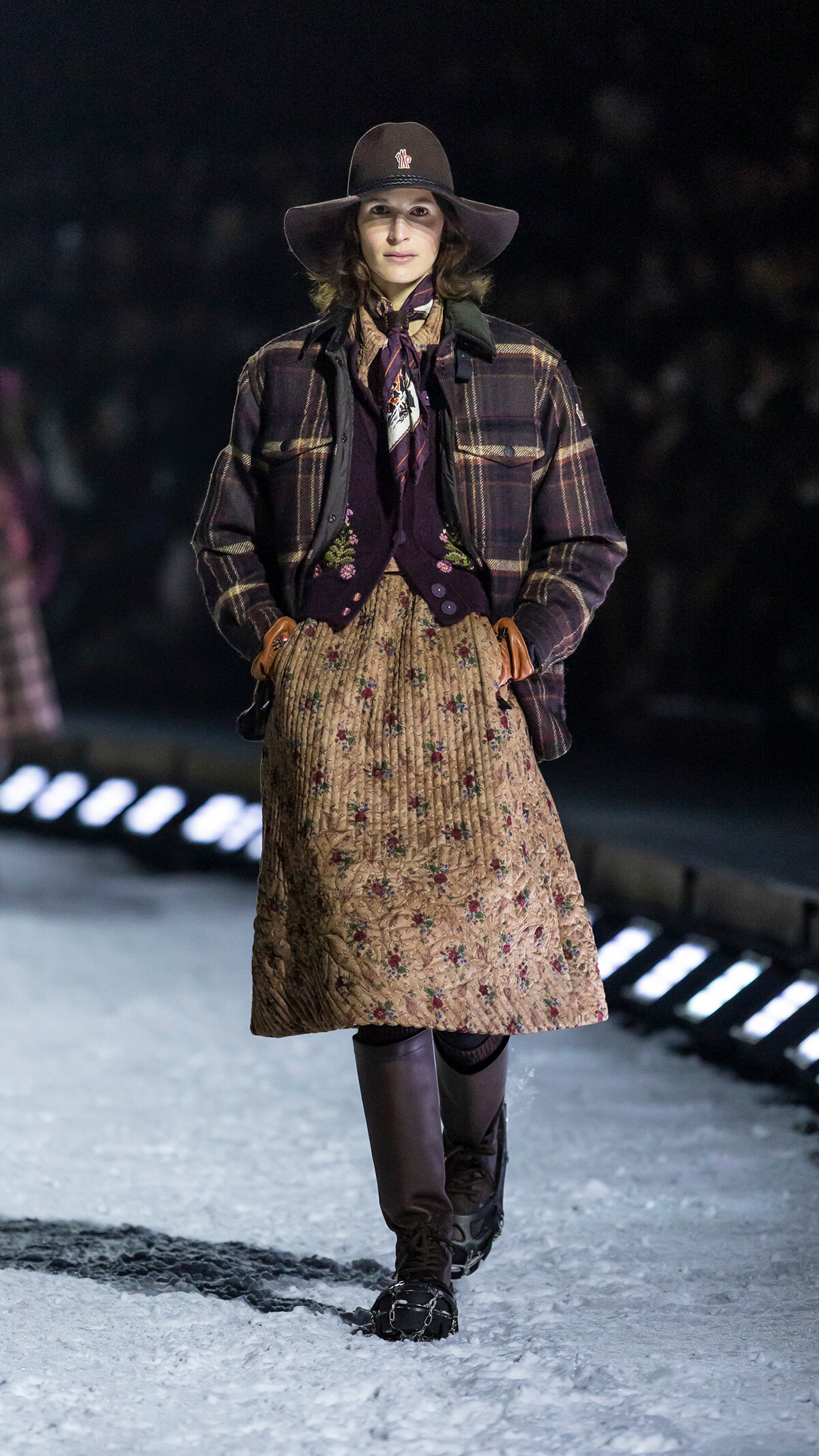 Model walking a snowy runway in a brown wide-brim hat, plaid jacket, floral midi skirt, and knee-high boots with brown gloves.