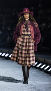 Female model walking a snowy runway in a burgundy plaid dress with a wide belt, scarf, and oversized cardigan.