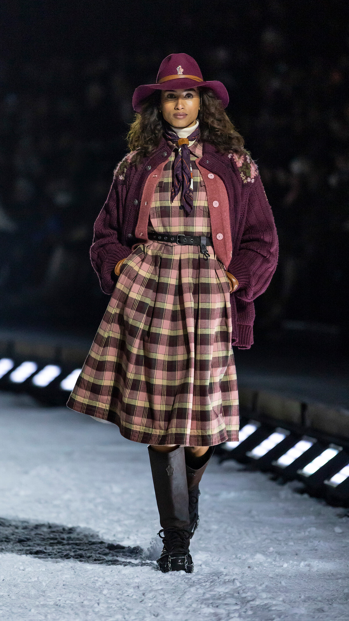 Female model walking a snowy runway in a burgundy plaid dress with a wide belt, scarf, and oversized cardigan.
