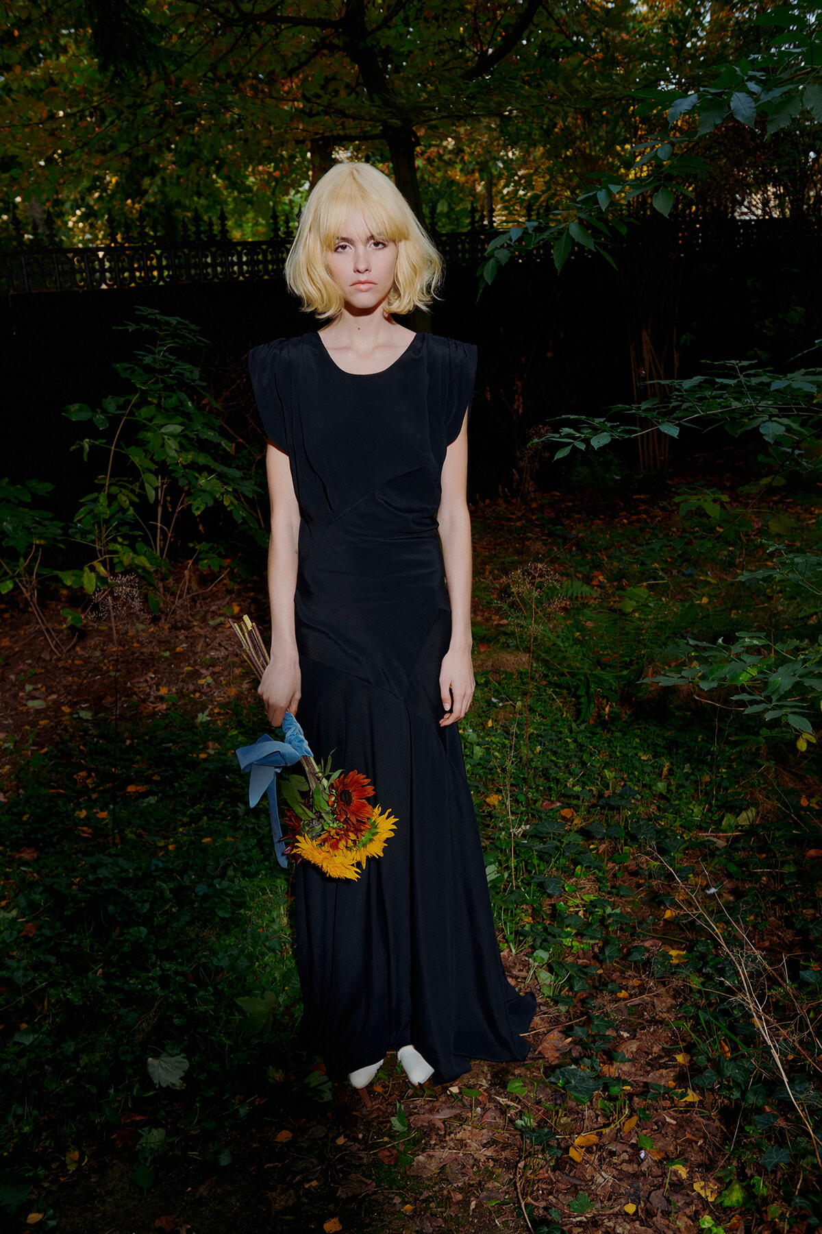 Woman in a black gown stands in a garden, holding a bouquet of sunflowers tied with a blue ribbon.