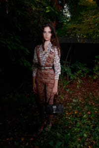 Young woman standing in a shadowy garden, wearing a burgundy patterned jumpsuit over a white printed blouse and a belt, holding a black handbag.