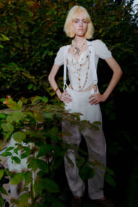 Young woman with short platinum blonde hair stands with hands on hips among dense green foliage, wearing an all-white, vintage-inspired outfit with layered pearl necklaces and suspenders.