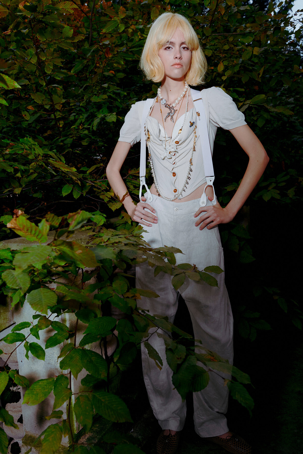 Young woman with short platinum blonde hair stands with hands on hips among dense green foliage, wearing an all-white, vintage-inspired outfit with layered pearl necklaces and suspenders.