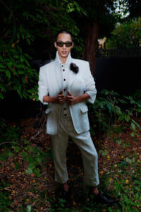 Man in a pale suit and dark sunglasses posing outdoors among green foliage.