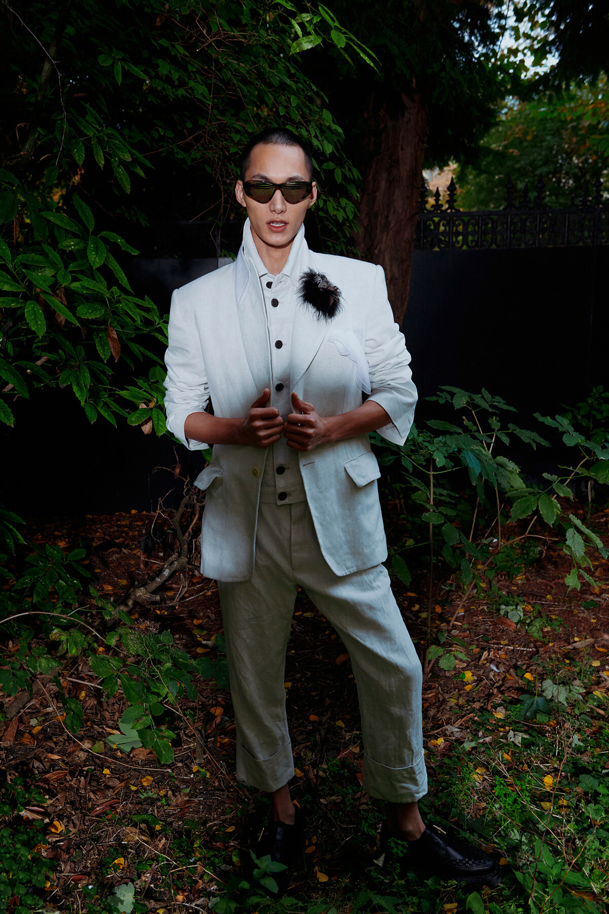 Man in a pale suit and dark sunglasses posing outdoors among green foliage.