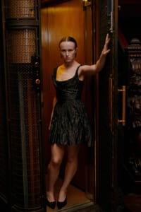 Woman in a black crinkled dress stands in an open elevator doorway, one hand against the wall and looking at the camera.
