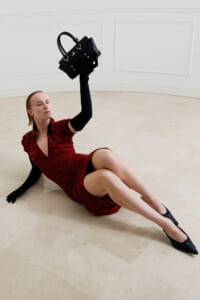 Model in a red textured dress sits on a beige tiled floor, raising a black handbag above her head with one gloved arm.