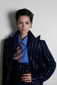 Fashion portrait of a person in a navy pinstripe suit, blue striped shirt, and red striped tie, posing against a light wall.