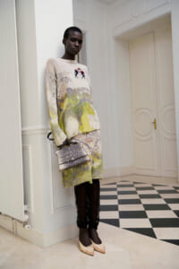 Model standing in a light hallway wearing a cream oversized knit dress with a pastel abstract print, black lace leggings, nude pointed heels, and a small gray patterned handbag.