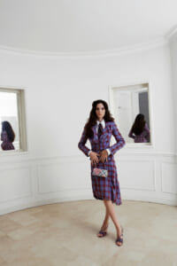 Woman in a red-blue plaid suit with white shirt and tie posing in a bright white room, holding a small plaid handbag.