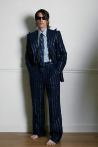 Man in a navy pinstripe suit and matching tie, standing barefoot against a white wall with hands in pockets and dark sunglasses on.