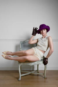 Woman in a cream lace top and shorts sits sideways on a vintage carved chair, wearing a purple hat and brown gloves.