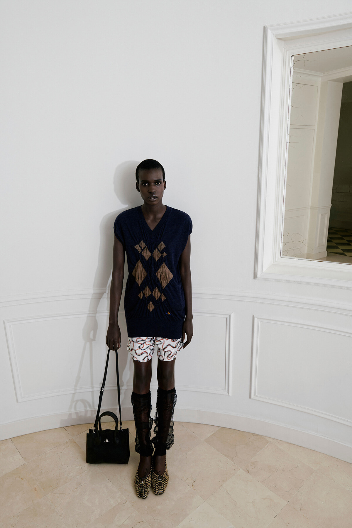 Young Black woman standing against a white paneled wall, wearing a navy V-neck sweater with brown argyle pattern, white patterned shorts, black lace knee-high socks, tan woven flats, and a small black handbag.