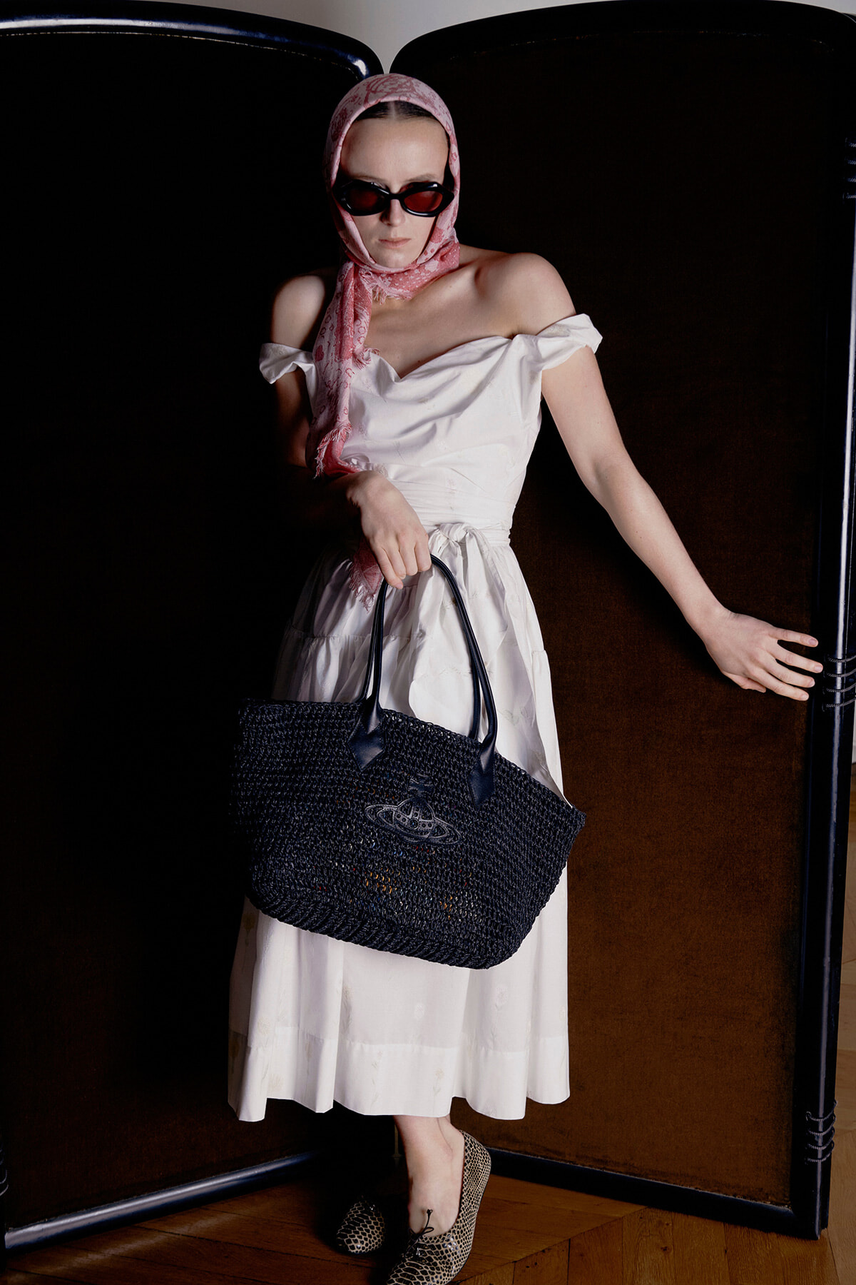 Woman in a white off-shoulder midi dress, pink headscarf and dark sunglasses, holding a large black woven tote as she leans against a dark divider.