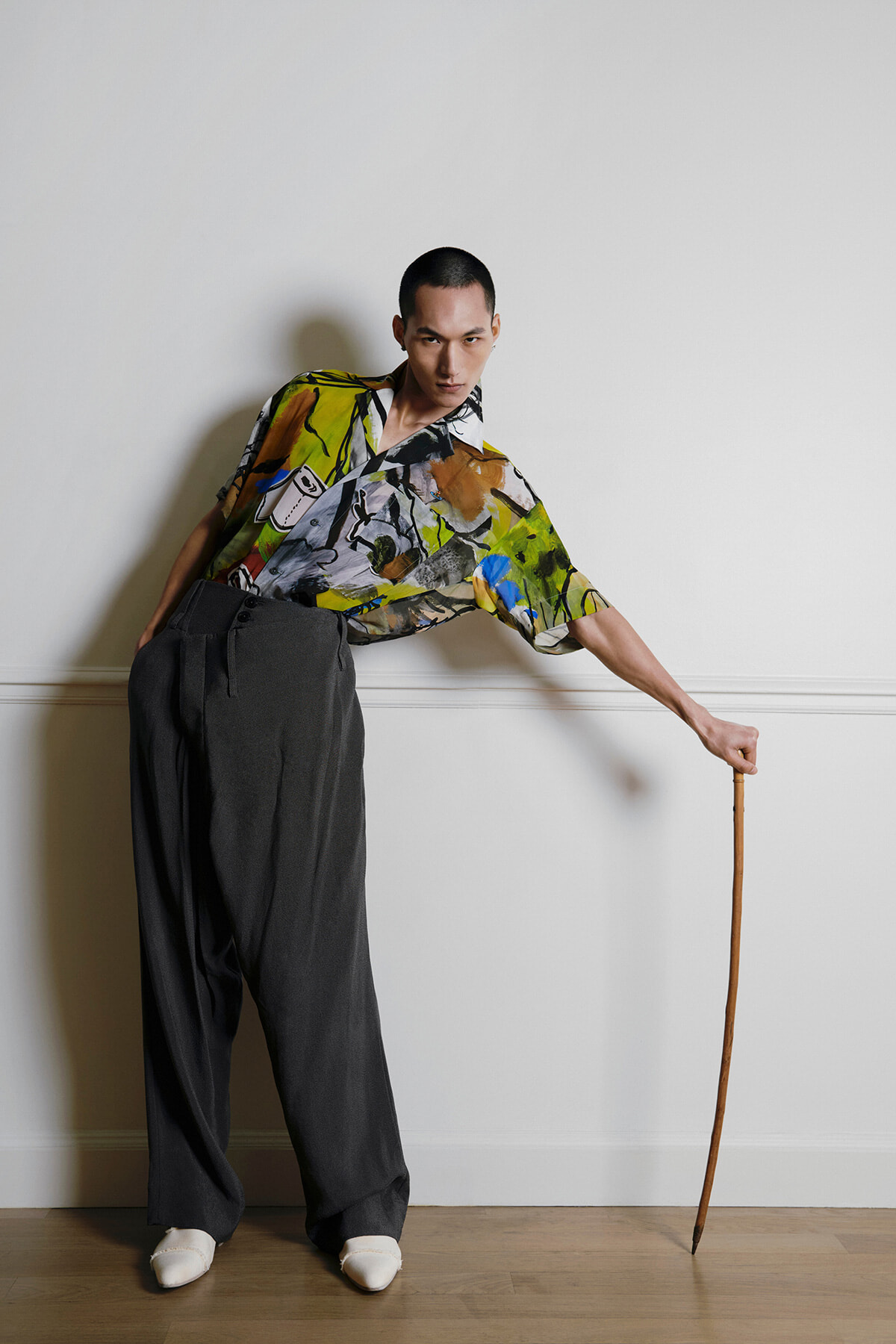 Person leaning to the side in a colorful abstract shirt and wide dark pants, holding a wooden cane indoors.