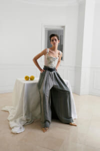 Fashion model stands with hands on hips in a bright white room, wearing a corset-style top and oversized pleated gray trousers, beside a table with two yellow apples.