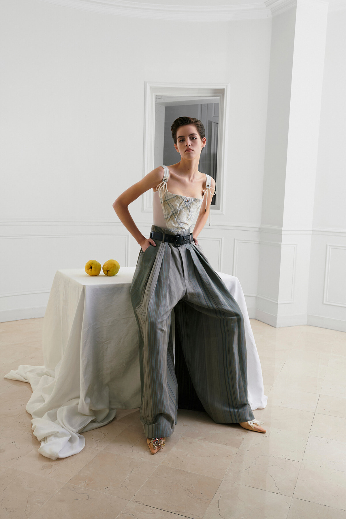 Fashion model stands with hands on hips in a bright white room, wearing a corset-style top and oversized pleated gray trousers, beside a table with two yellow apples.