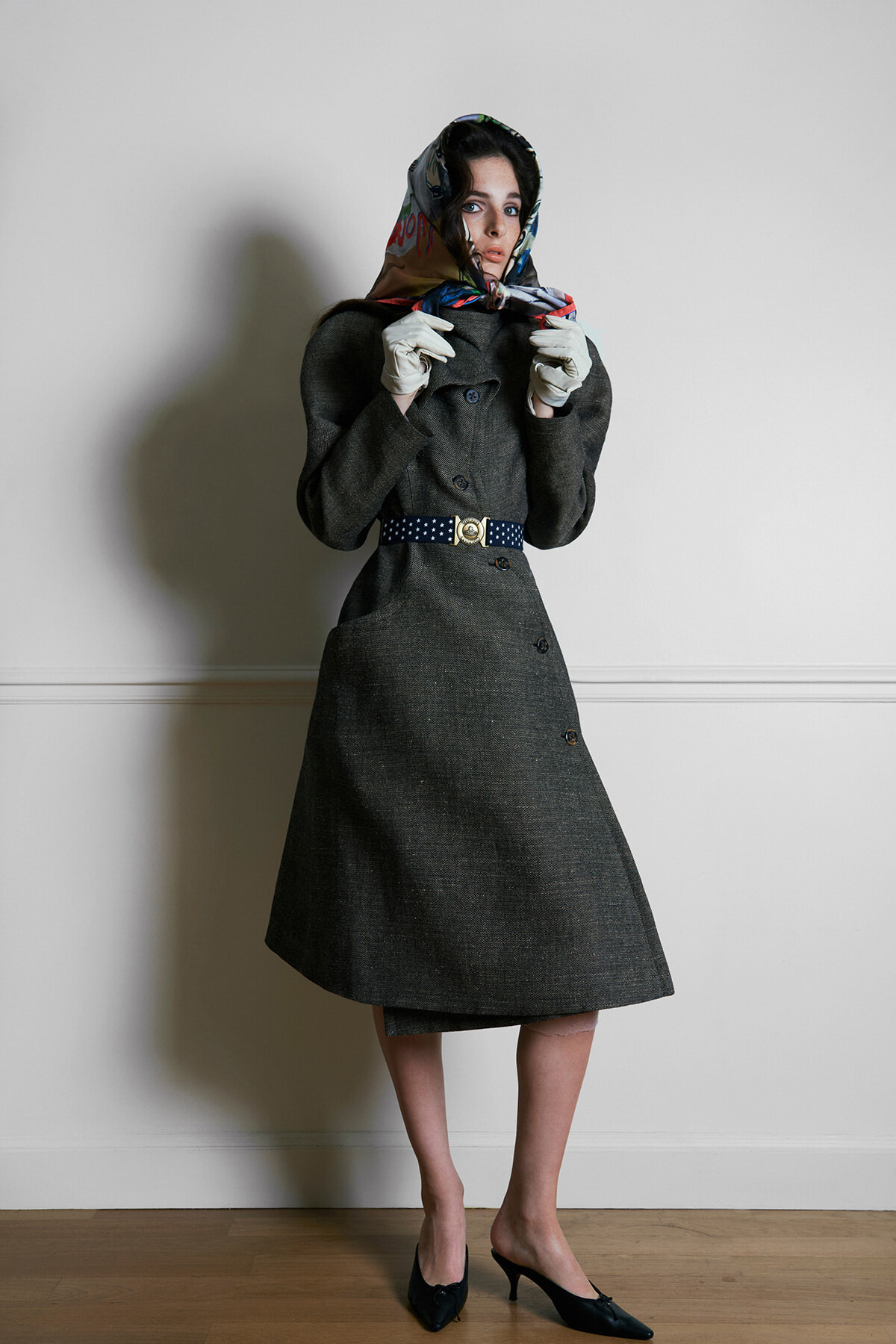 Fashion portrait of a woman in a dark mid-century coat with a patterned scarf wrapped around her head and white gloves, standing indoors against a pale wall with hands near her collar.