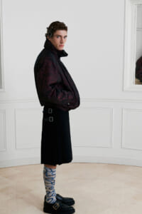 Man in a dark patterned quilted jacket and black midi skirt with buckles, blue patterned socks, and loafers, posed sideways in a white room.