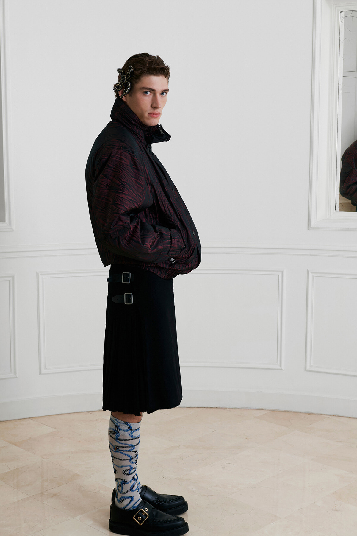Man in a dark patterned quilted jacket and black midi skirt with buckles, blue patterned socks, and loafers, posed sideways in a white room.