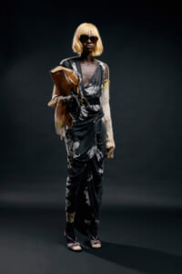 Model in a dark floral wrap dress with a belt, blonde bob, oversized sunglasses, carrying a tan handbag and scarf.