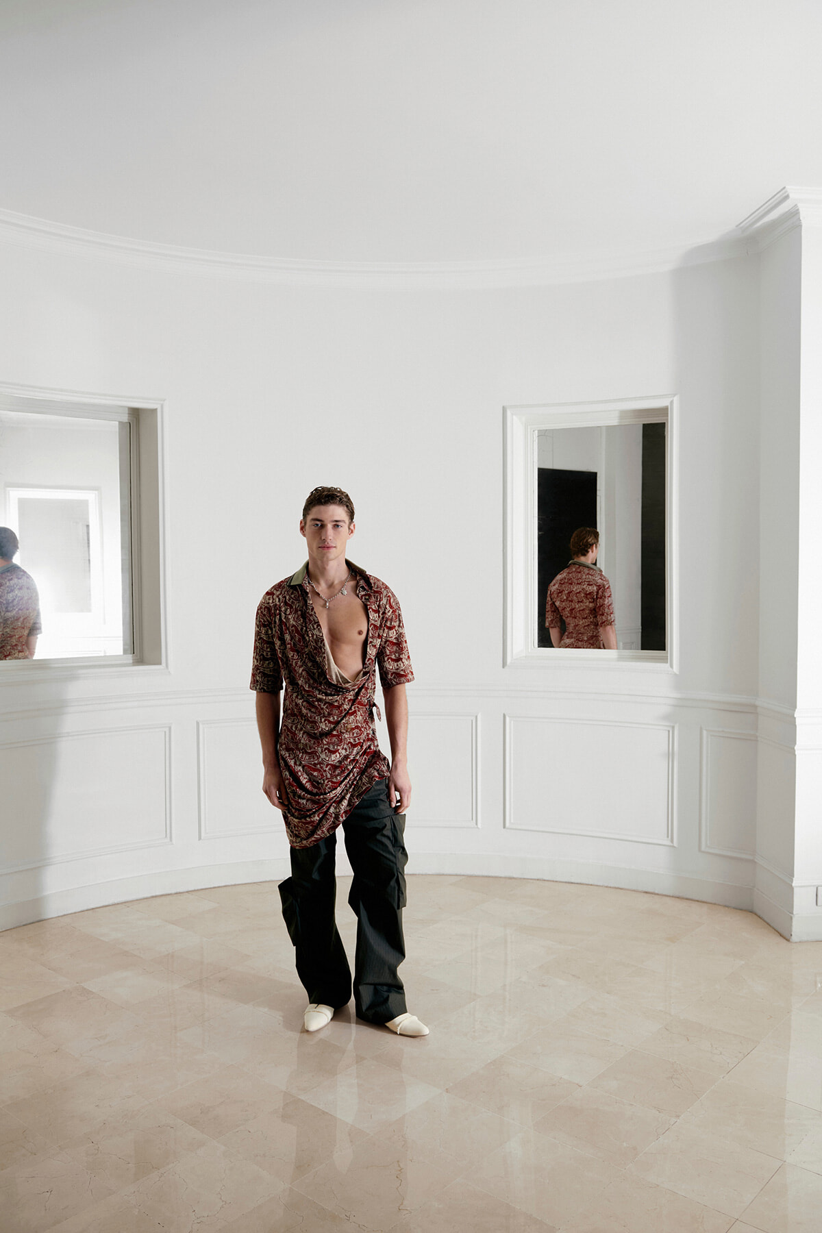 Young man in a red patterned, open shirt and black cargo pants stands in a bright white curved-room with mirrors showing his reflection.