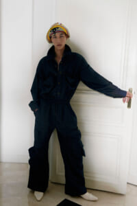 Person in a dark jumpsuit leaning against a white door, wearing a colorful striped headwrap and light-colored shoes.