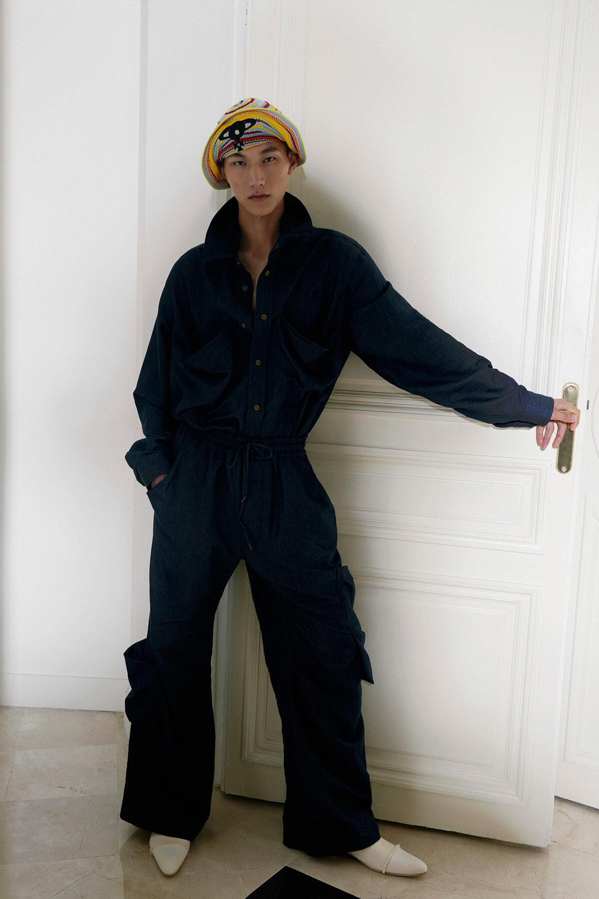 Person in a dark jumpsuit leaning against a white door, wearing a colorful striped headwrap and light-colored shoes.