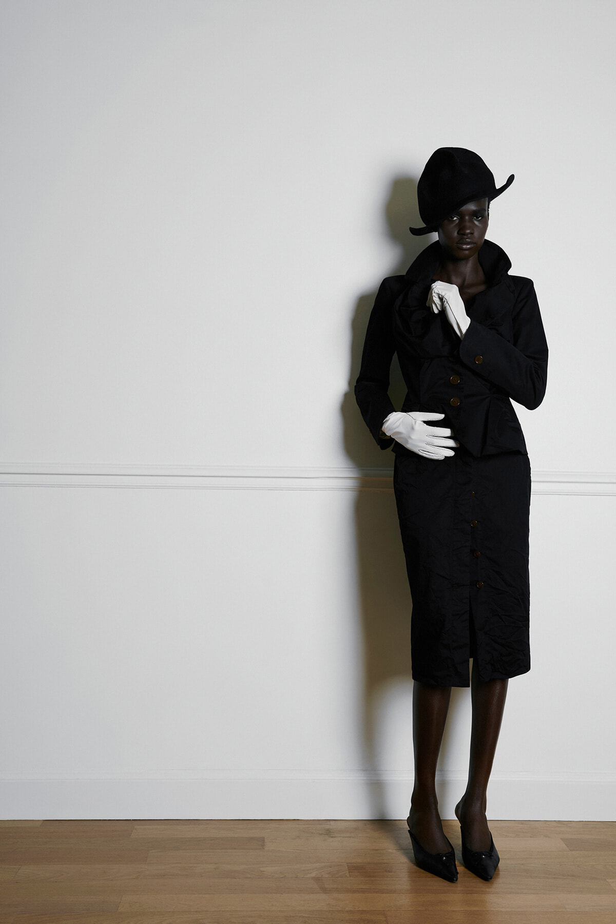 Woman in a black coat, matching hat, white gloves, and black heels posing against a plain white wall.