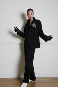 Male model in a black suit with a decorative brooch, white pocket square, and black gloves posing against a pale wall in a fashion shoot.