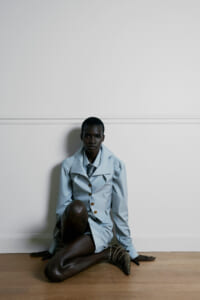 Dark-skinned person sits on a wooden floor against a white wall, wearing a light blue blazer and matching shorts, with animal-print boots nearby.