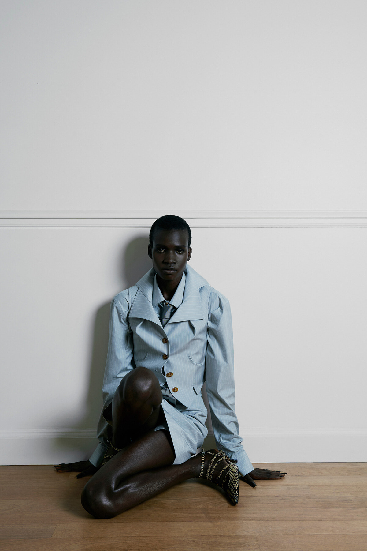 Dark-skinned person sits on a wooden floor against a white wall, wearing a light blue blazer and matching shorts, with animal-print boots nearby.