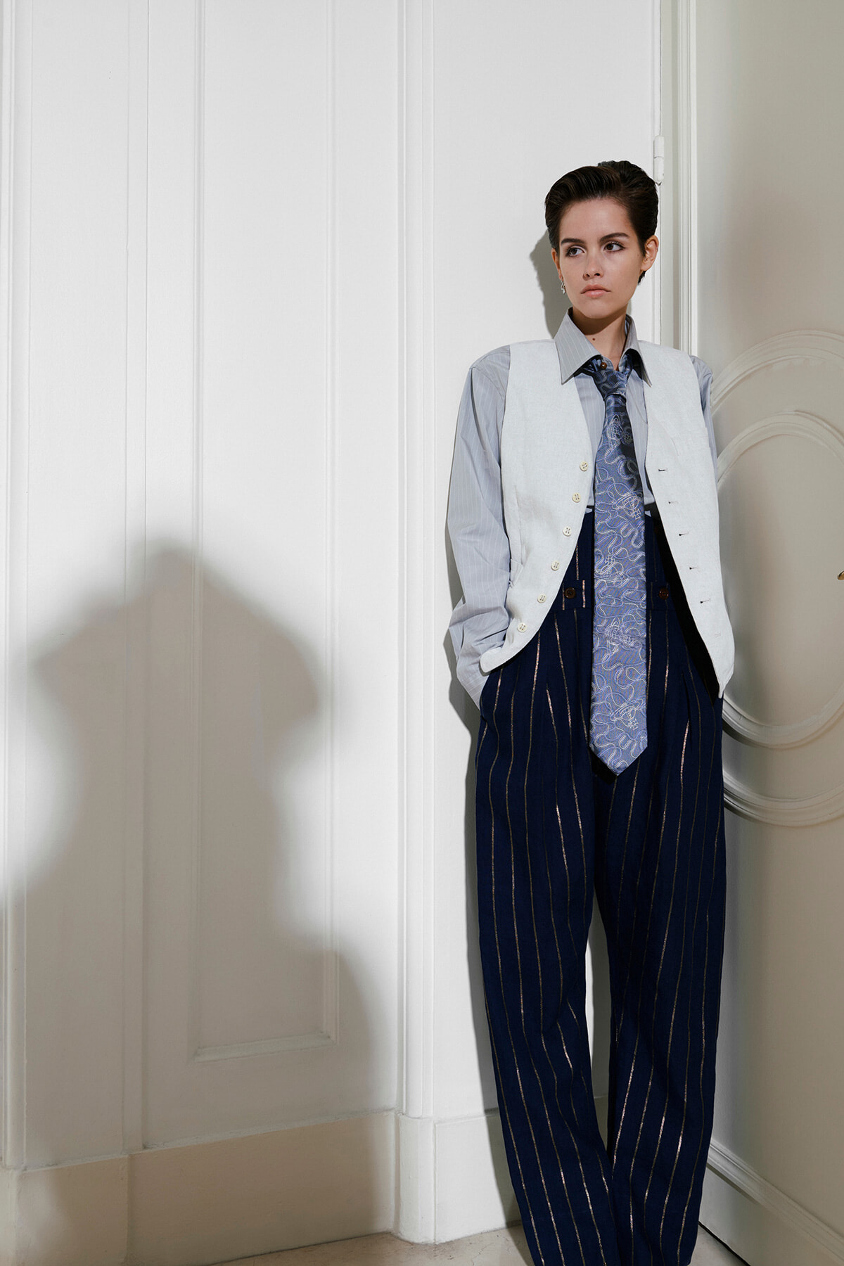 Person standing in a doorway wearing a light vest over a blue shirt, and dark pinstriped trousers with a patterned tie.