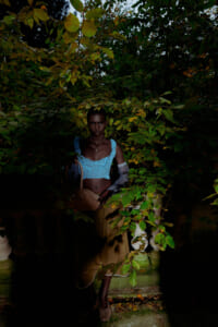 Woman wearing a blue lace crop top and beige skirt stands among dense green foliage, holding a tan handbag at dusk.