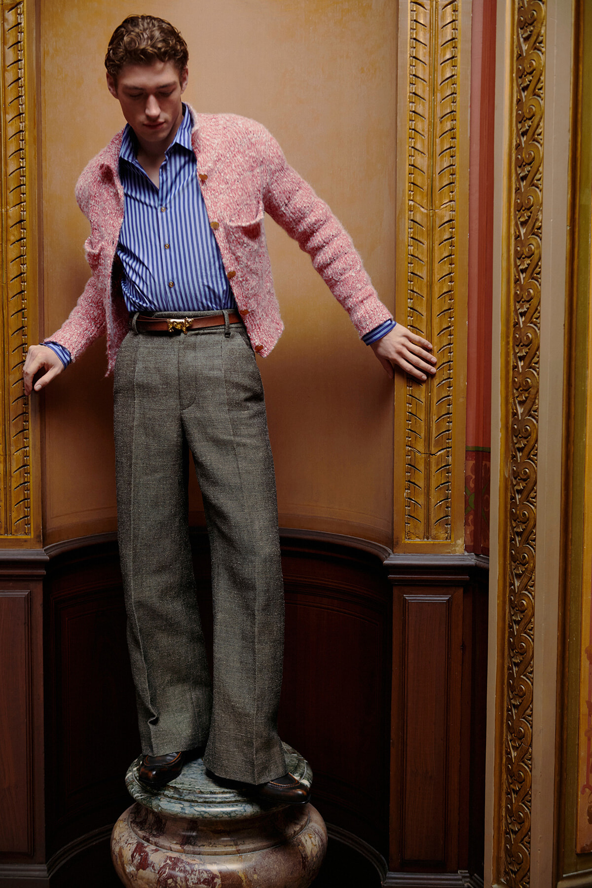 Man in a pink knit cardigan, blue striped shirt, and gray wide-leg trousers stands on a marble pedestal in an ornate, gold-toned interior.