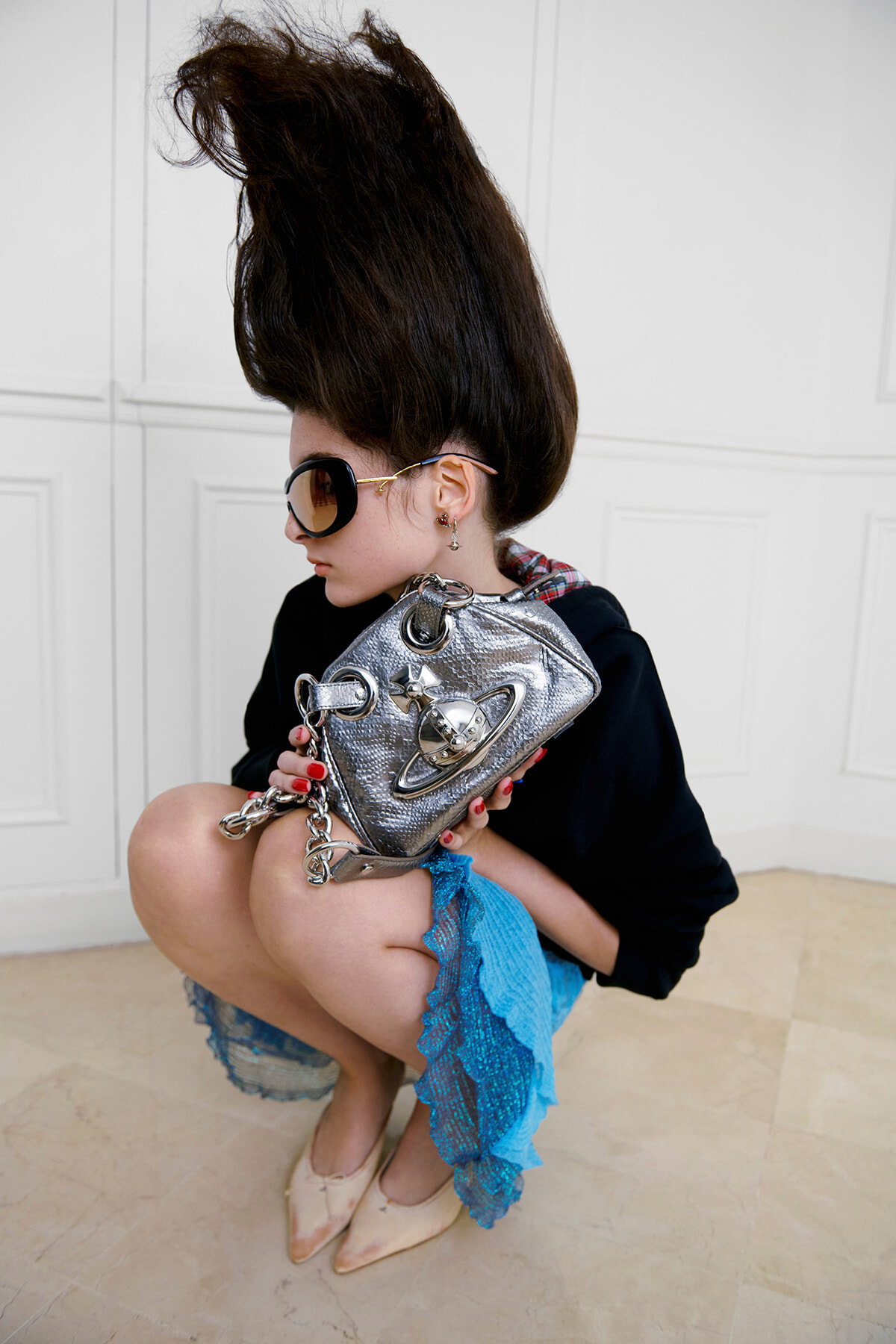 Woman squatting in a white room, wearing oversized sunglasses, a black top, blue lace skirt, beige heels, and a metallic silver handbag.