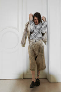 Person standing in a white paneled doorway, wearing a gray patterned sweater, a light denim jacket draped over shoulders, and tan cropped wide-leg pants; visible tattoo on lower leg; black loafers.