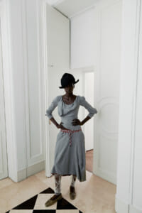 Woman in a grey dress with a red belt and lace leggings stands with hands on hips in a bright white room.