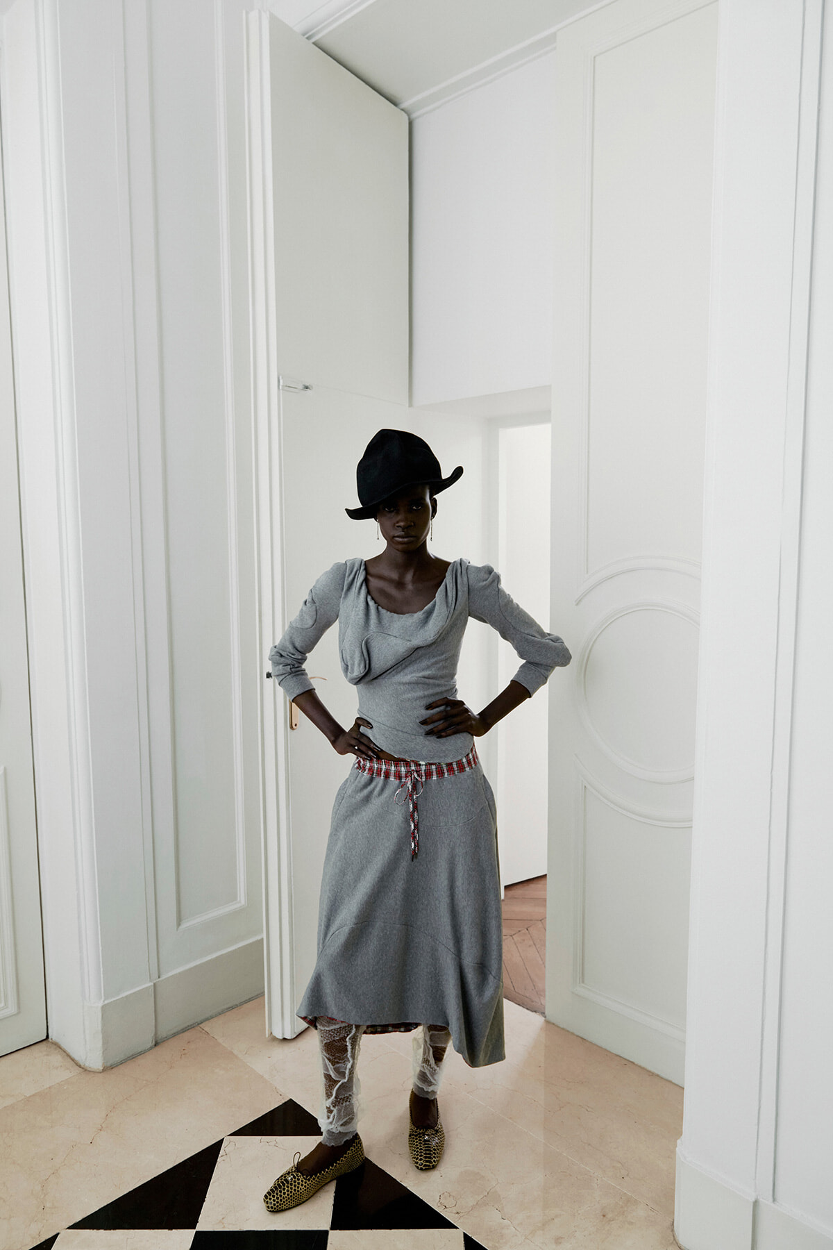 Woman in a grey dress with a red belt and lace leggings stands with hands on hips in a bright white room.